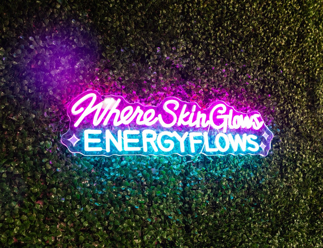 Where Skin Glows, Energy Flows neon sign on the studio greenery wall
